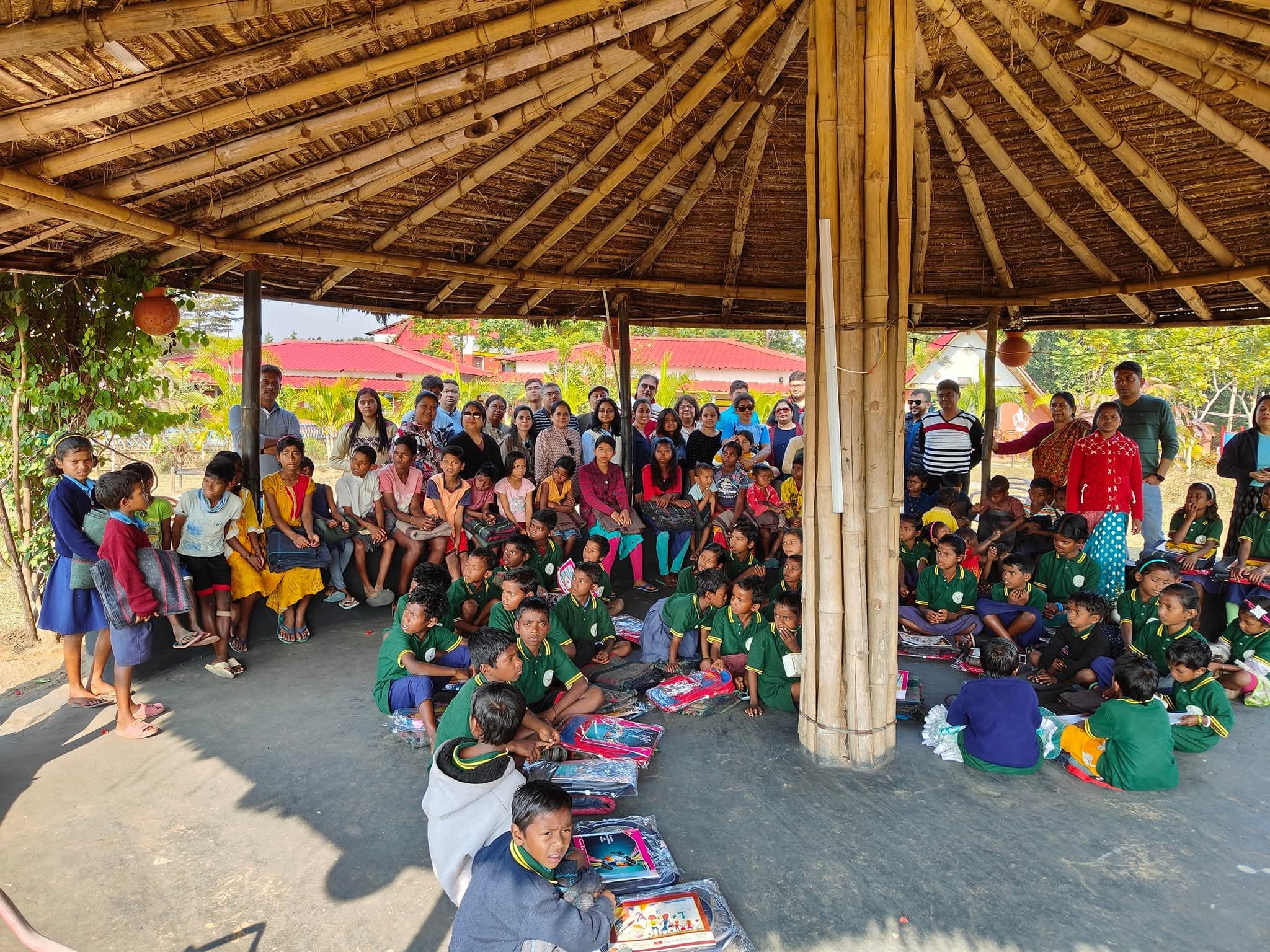 Large group of children and teachers gathered under a pavilion with books being distributed